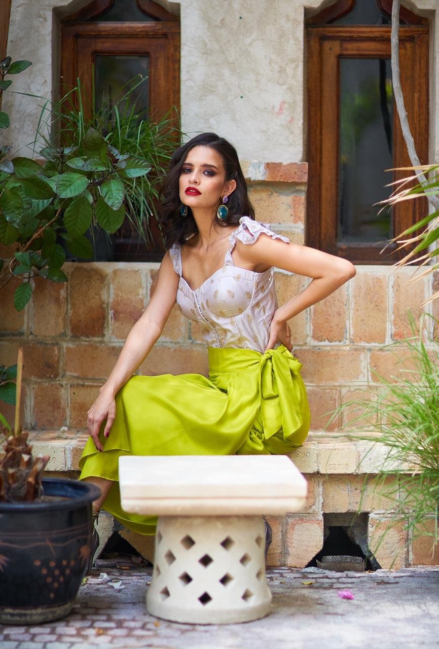 Woman in a white top and green skirt sitting on a stone bench with plants around.