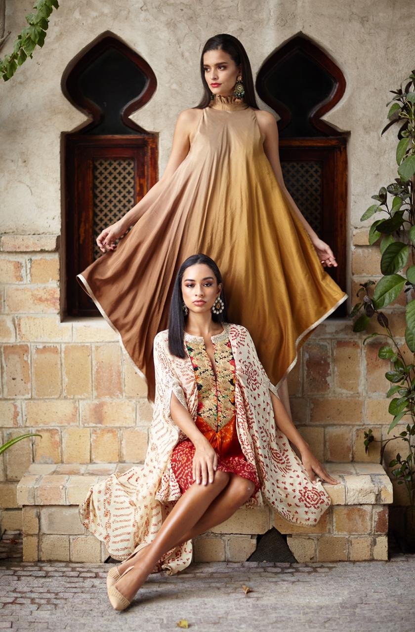 Two women in traditional outfits sitting on a stone step with a textured wall and plants in the background.