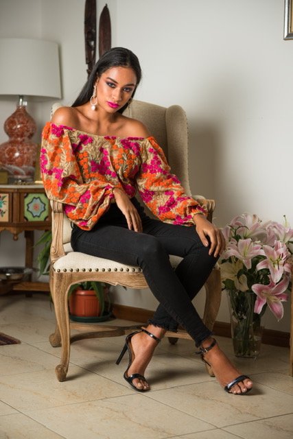 Woman in a colorful off-shoulder top sitting on a chair in a room with flowers and decor.