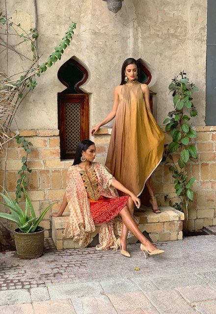 Two women in colorful dresses standing in a rustic outdoor setting with plants and stone walls.