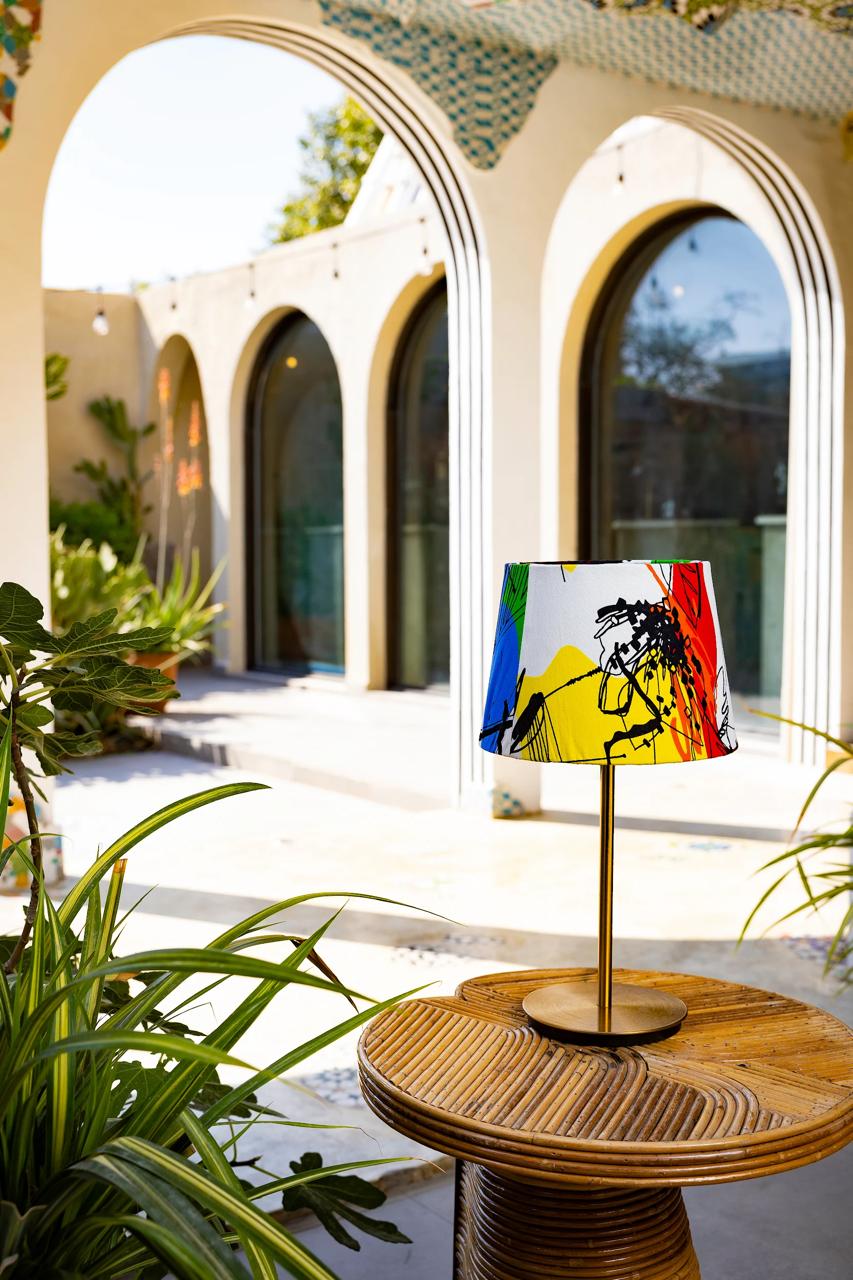 Decorative lamp with a colorful shade on a wooden table in an outdoor setting with arches and plants.